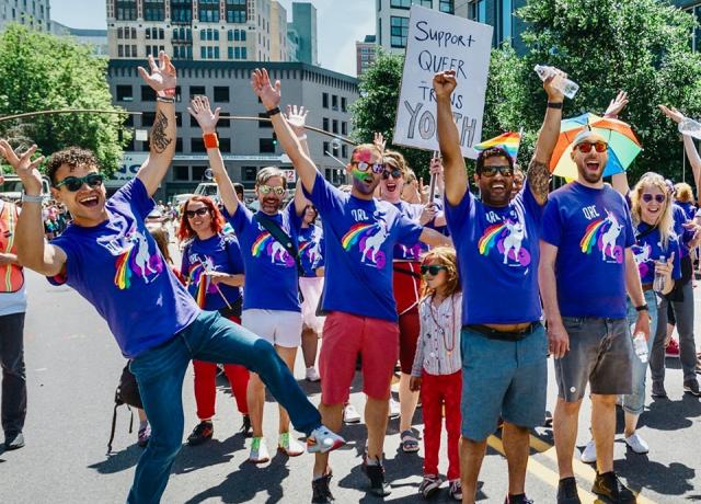 a group of people all wearing purple QRC shirts with a unicorn puking a rainbow stand outside happily with their arms raised in celebration at pride